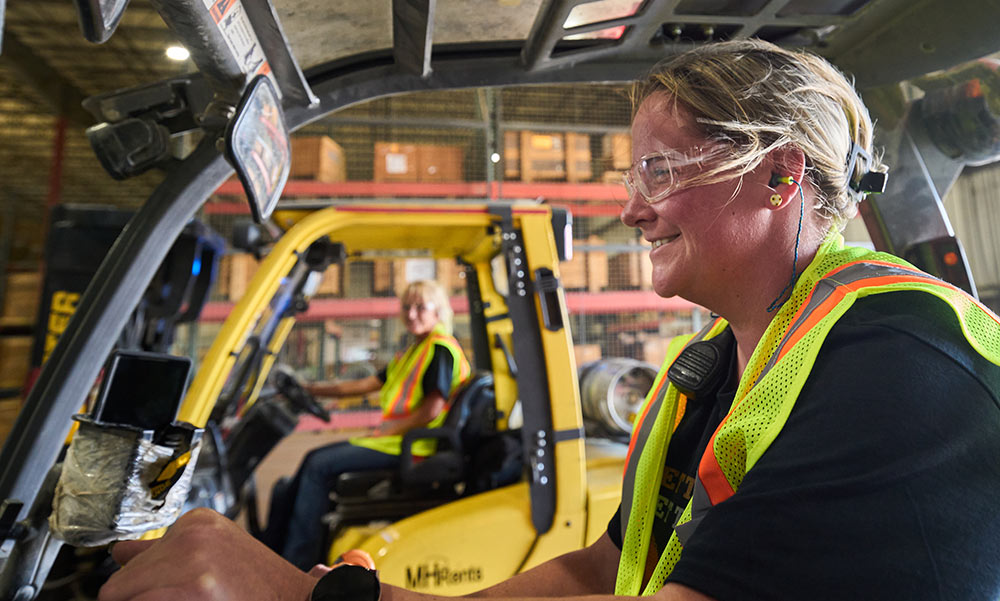 A female forklift operator smiles as she drives by in a warehouse